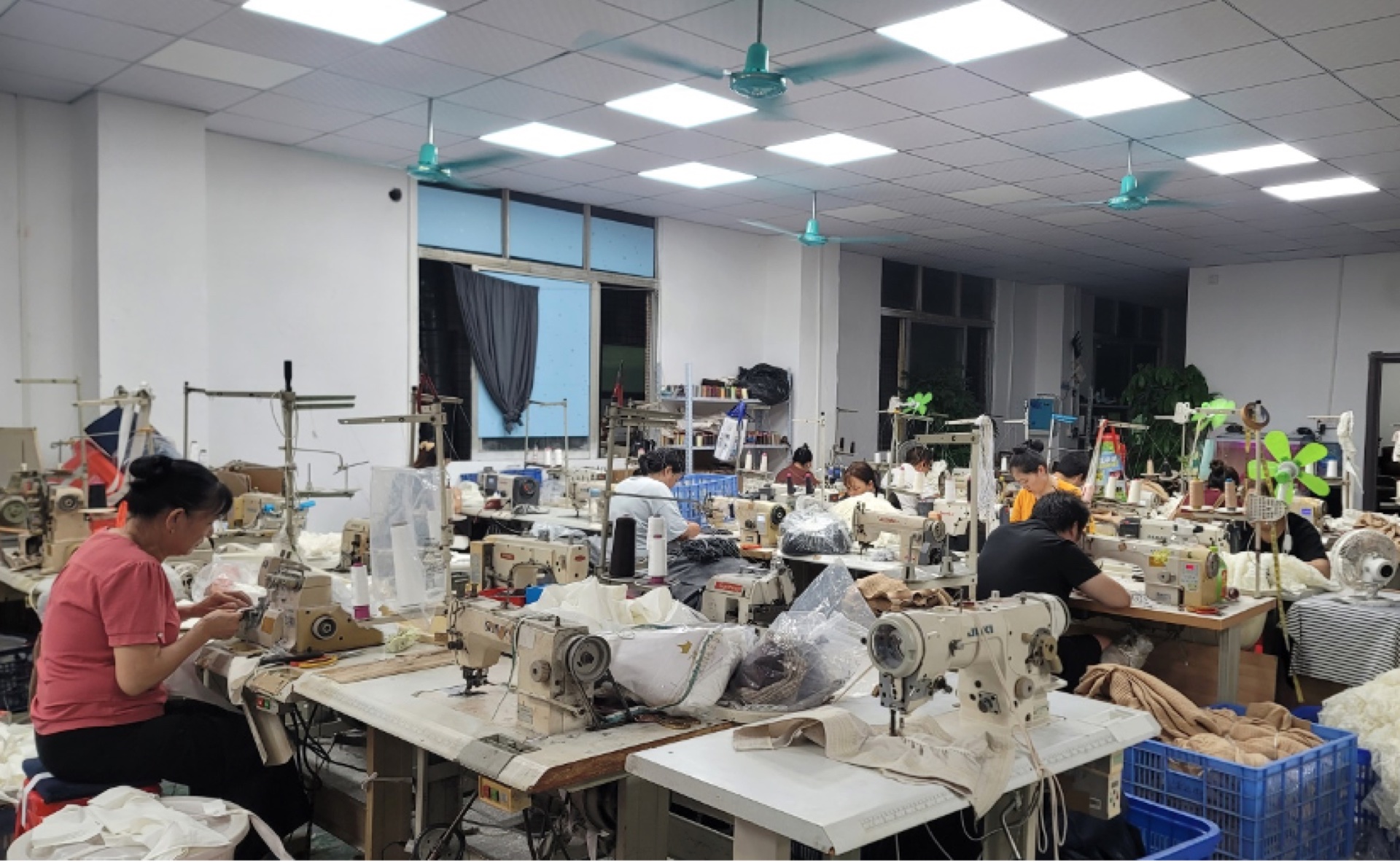 Garment workshop with workers sewing at industrial machines along long workbenches.