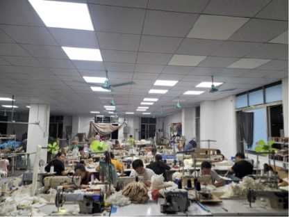 Garment production workshop with staff working at long tables with fabrics, patterns, and sewing areas in the background.