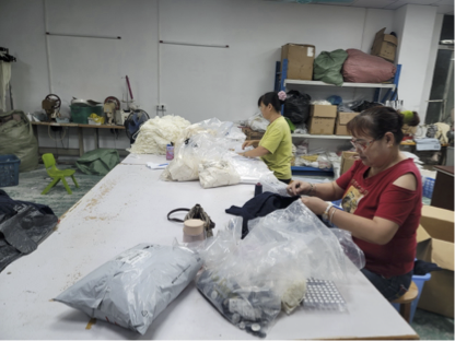Two workers at a large table in a garment workshop with textile materials, yarn, and shipping packages.